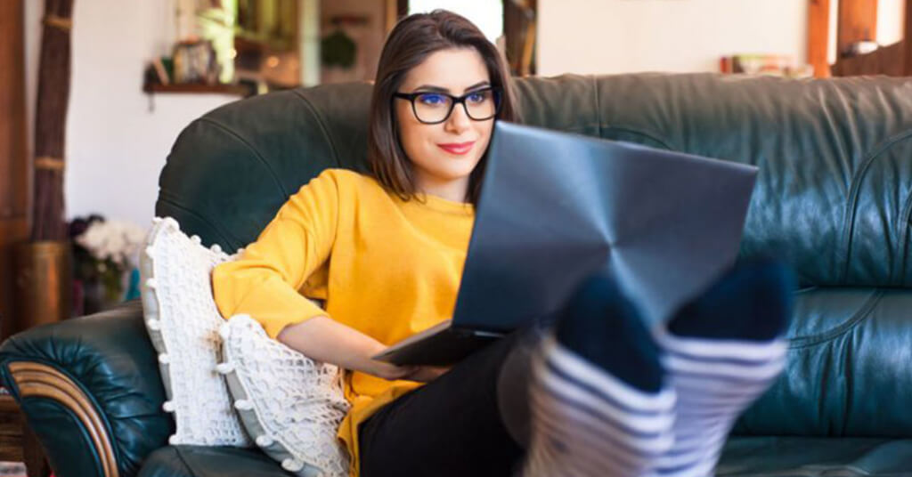 woman in yellow sweater, glasses, sitting on couch on computer